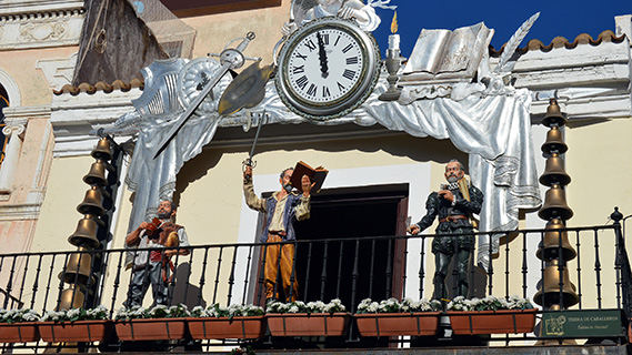 Eine Uhr mit weißem Zifferblatt krönt einen schmiedeeisernen Balkon, der von Säulen mit Bronzeglocken flankiert wird