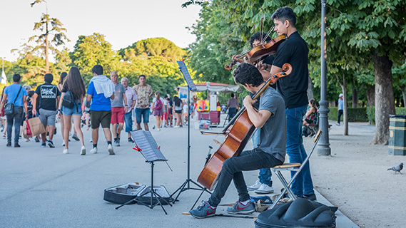 Drei junge Menschen – zwei an der Geige und einer am Cello – spielen auf einer weitläufigen, von Bäumen gesäumten Promenade im Retiro-Park in Madrid