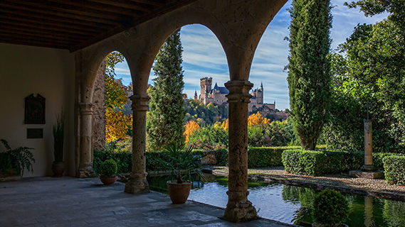 Hinter Steinbögen eröffnet sich ein Teich mit ruhigem Wasser, im Hintergrund ist der Alcázar von Segovia zu sehen