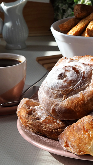 Mehrere goldbraune, spiralförmige und mit Puderzucker bestäubte Einsaimadas auf einem Teller neben einer Tasse heißer Schokolade