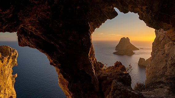 Durch einen natürlichen Bogen aus rauem, orangefarbenem Kalkstein lässt sich bei Sonnenuntergang die pyramidenförmige Silhouette einer kleinen Insel erkennen