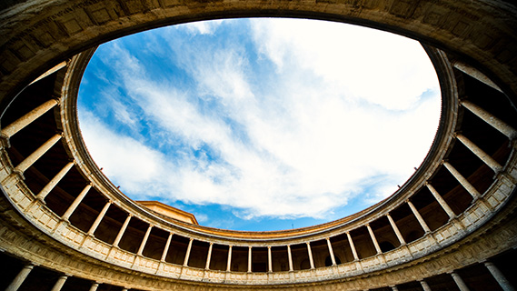 Aufnahme aus der Froschperspektive des blauen Himmels mit weißen Wolken durch das riesige Rundfenster, das den Renaissance-Innenhof des Palasts von Karl V. umrahmt