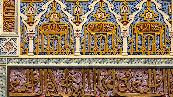 Eine Nahaufnahme einer kunstvollen Stuckverzierung, bei der sich vergoldete arabische Schriftzeichen mit Pflanzenmotiven vor blauem und rötlichem Hintergrund verflechten